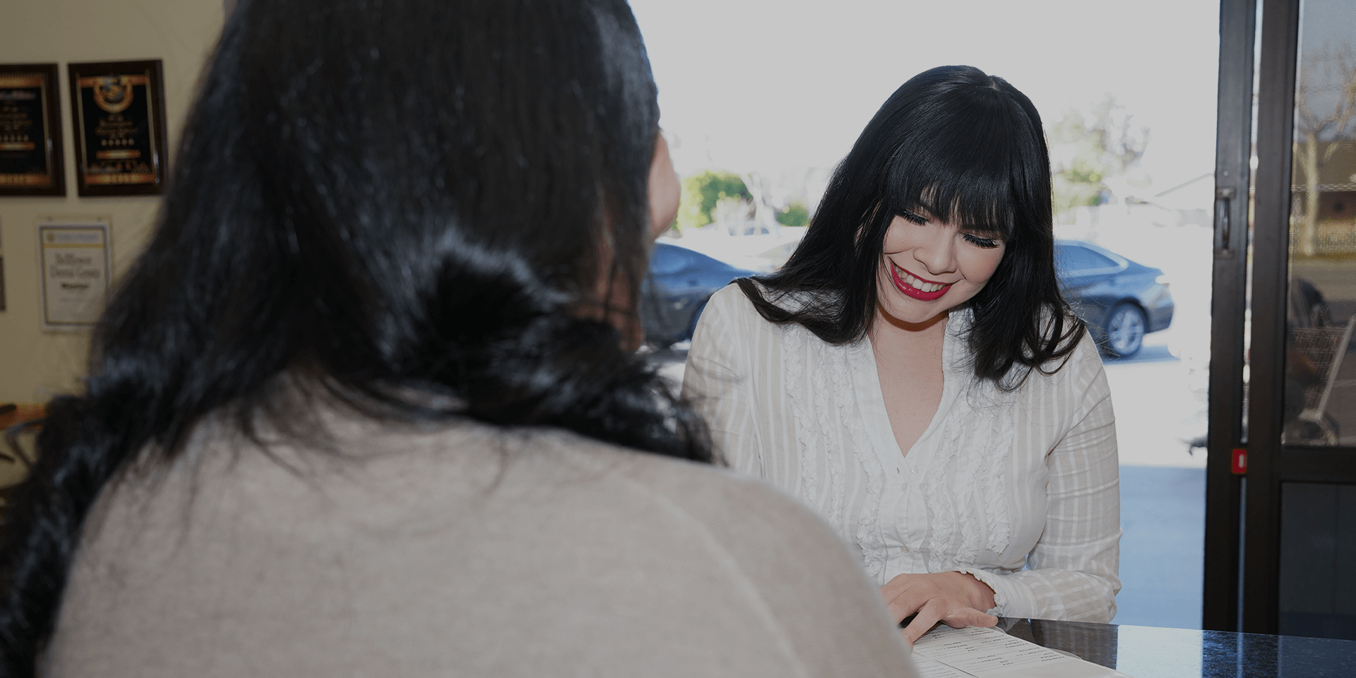 A woman smiling while signing paperwork with a front desk staff member.