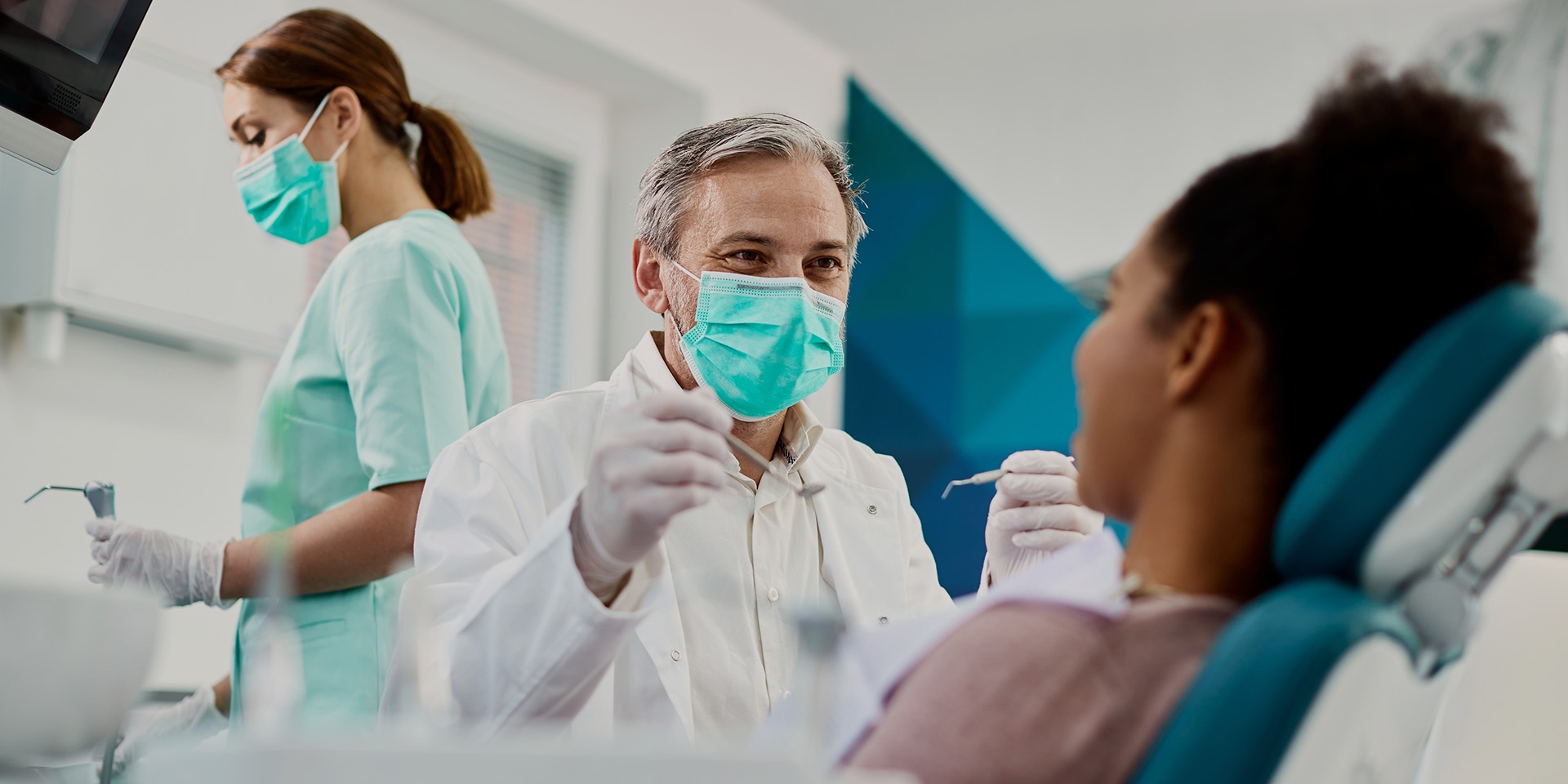 A dentist wearing a mask holding dental tools while a patient is seated in the dental chair.