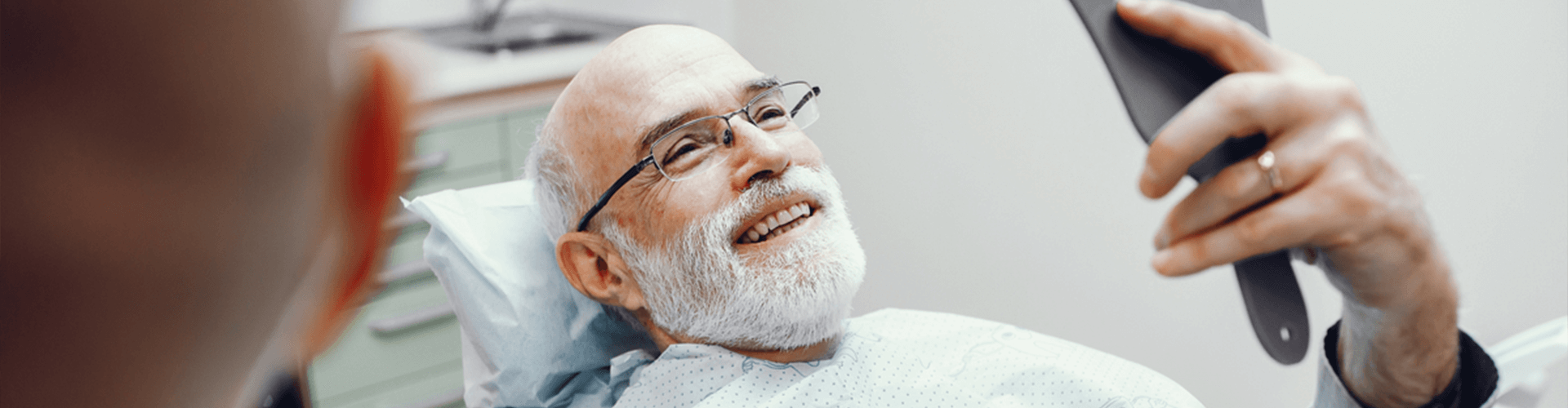Man in dental chair smiling in hand mirror.