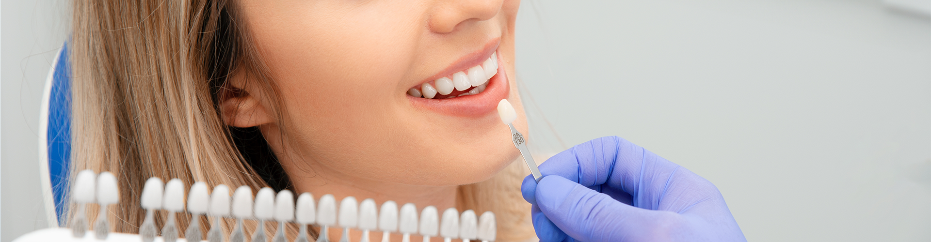 gloved hands holding tooth shade-matching tool against woman's smile