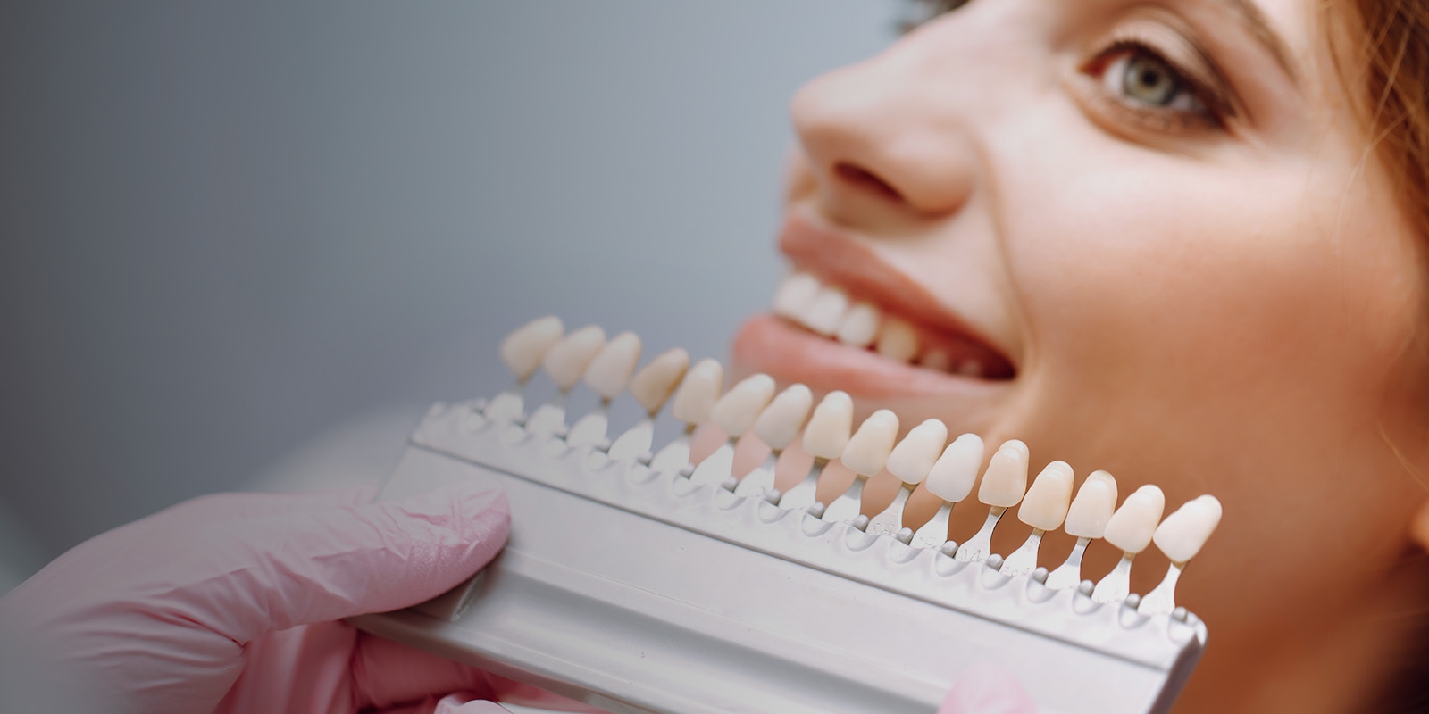 A woman smiling with a dental veneer sample held in front of her teeth.