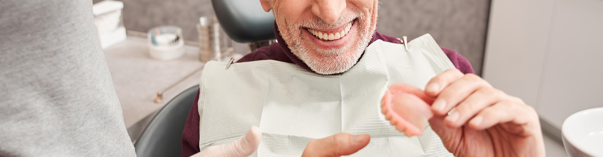 smiling man holding upper denture in dentist chair