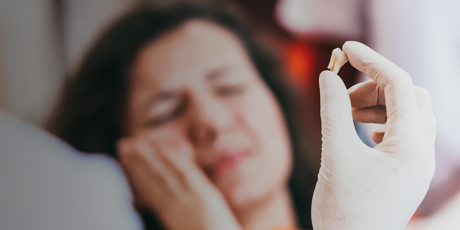 A white-gloved hand holding a tooth with a patient in the background showing a discomforted expression.